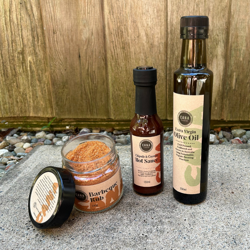 Collection of barbecue products including a bottle of olive oil, hot sauce, and barbecue rub on a stone surface with a wooden background.